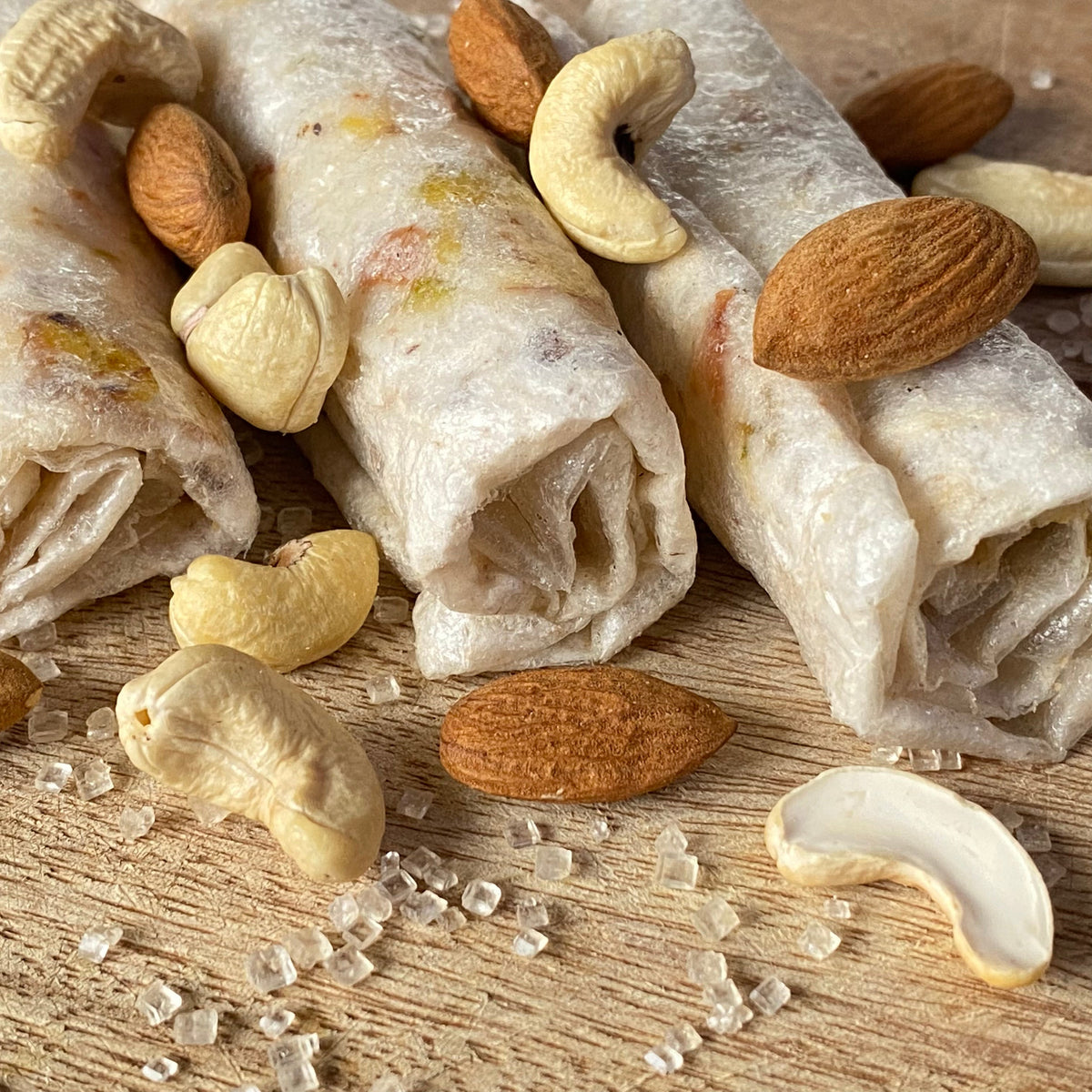 Close-up of Sugar Kaju Badam Pootharekulu rolls surrounded by almonds, cashews and raw sugar crystals showing the paper-thin sweet texture